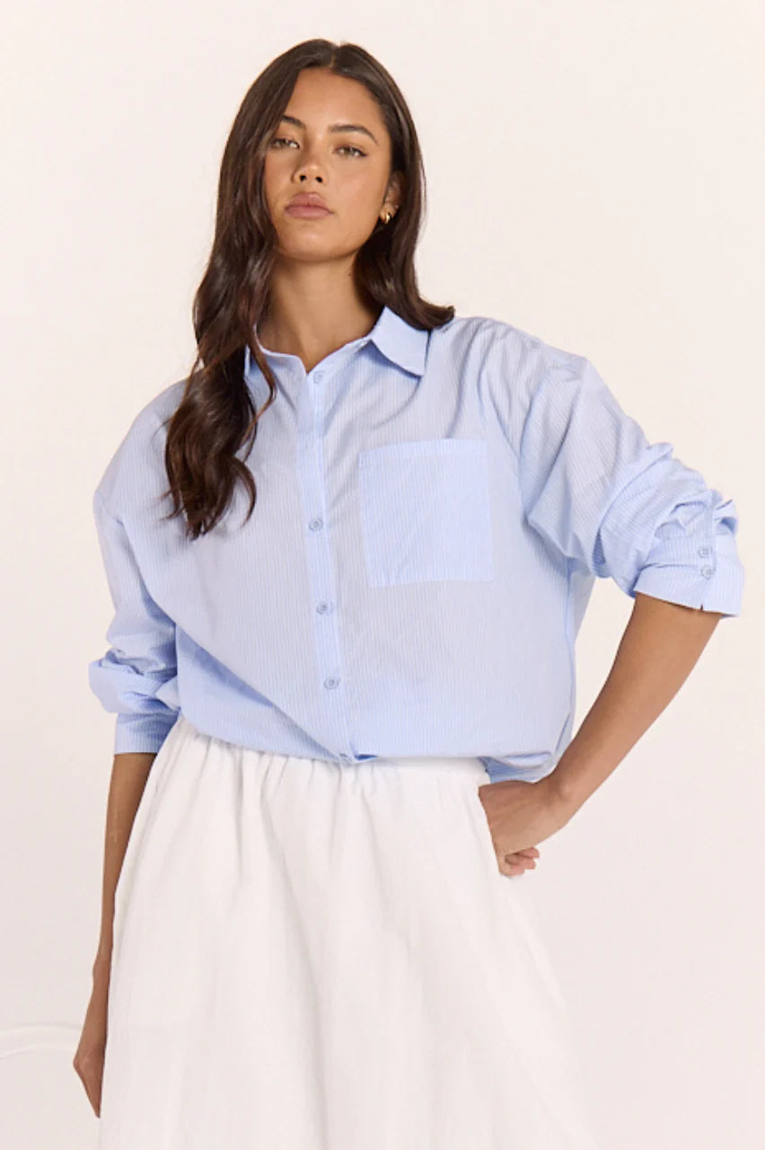 Blue striped Emily Shirt by Feather + Noise, shown hanging on a wooden hanger with a relaxed fit and classic collar.